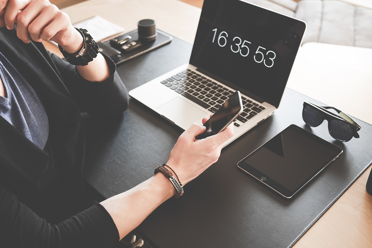 Das Bild zeigt einen Laptop-Bildschirm mit Zeitangabe und eine Person, die mit einem Smartphone in der Hand davorsitzt